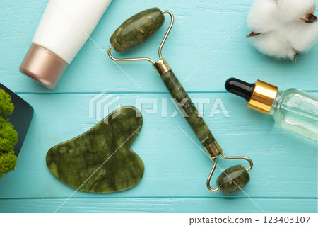 Green jade roller and gua sha stone for facial massage and on blue background. Home beauty and selfcare accessories. Face roller for anti age wrinkle treatment. Top view, flat lay. 123403107