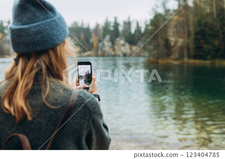 Person in hat is recording content with a smartphone near lake on wild nature background. Stylish 30s girl with phone walking outdoors. Travel, adventure. Concept of an active lifestyle. 123404785