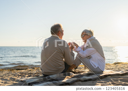 Senior mature couple sitting on beach holding hands enjoying outdoor recreation. Old husband wife touching hands with tenderness love. Grandmother grandfather together. Family moment of love 123405816