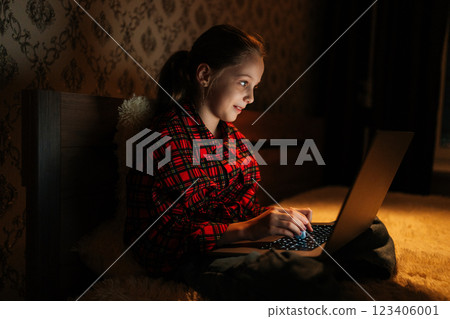 Side view of happy pretty child girl 10 yo with joyful expression using laptop sitting on bed in dark bedroom at night, warm lighting creating cozy and intimate atmosphere, illuminated by screen light 123406001