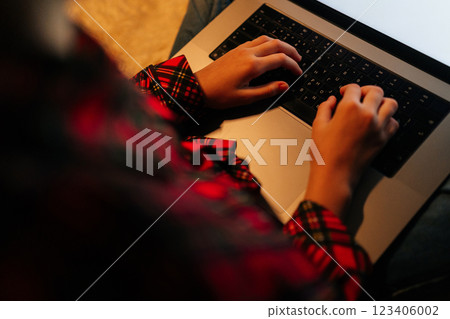 View from shoulder of unrecognizable child girl wearing red and black checkered shirt typing on backlit laptop keyboard sitting on bed with crossed legs in dark room, creating cozy atmosphere. 123406002