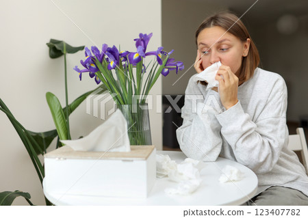 Sick unhealthy allergic young woman covering nose with paper tissue has runny stuffy nose, sneezes from iris flowers pollen at kitchen home. Girl with flu, itching or cough, seasonal allergy,rhinitis. 123407782