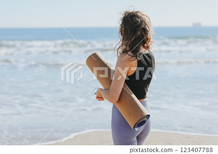 Rear view of slim lady in sportswear holds yoga mat and prepares to workout on sea beach. Sport fitness outdoors. Healthy lifestyle, morning exercise, female wellness and power, healthcare, activities Rear view of slim lady in sportswear holds yoga mat and prepares to workout on sea beach. Sport fitness outdoors. Healthy lifestyle, morning exercise, female wellness and power, healthcare, activities 123407788