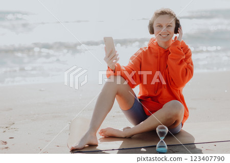 Workout playlist. Happy young caucasian woman with earphones sits on yoga mat outdoors on sea beach, listens to music or browsing internet with smartphone while resting after fitness exercising. Workout playlist. Happy young caucasian woman with earphones sits on yoga mat outdoors on sea beach, listens to music or browsing internet with smartphone while resting after fitness exercising. 123407790