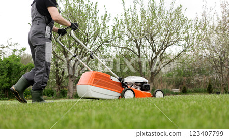 Professional gardener in protective apparel is mowing green grass lawn using modern gasoline cordless lawnmower at backyard. Seasonal landscaping design work. Blooming trees on background Professional gardener in protective apparel is mowing green grass lawn using modern gasoline cordless lawnmower at backyard. Seasonal landscaping design work. Blooming trees on background 123407799