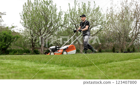 Professional gardener in protective apparel is mowing green grass lawn using modern gasoline cordless lawnmower at backyard. Seasonal landscaping design work. Blooming trees on background Professional gardener in protective apparel is mowing green grass lawn using modern gasoline cordless lawnmower at backyard. Seasonal landscaping design work. Blooming trees on background 123407800