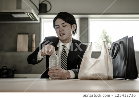 An Asian businessman in a suit drinks mineral water at the dining room after returning home An Asian businessman in a suit drinks mineral water at the dining room after returning home 123412736