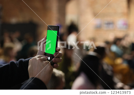 A person is holding a smartphone that features a green screen, surrounded by a vibrant crowd scene A person is holding a smartphone that features a green screen, surrounded by a vibrant crowd scene 123413973