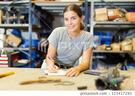 Smiling young woman storehouse worker standing at workbench and making notes Smiling young woman storehouse worker standing at workbench and making notes 123417293