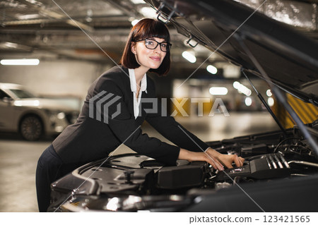 Confident Caucasian businesswoman in her 30s inspects car engine under hood in underground garage. Professional attire and focused expression convey self-reliance and determination. 123421565