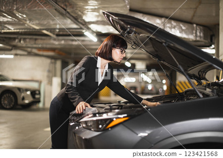 Caucasian businesswoman inspecting car engine in underground parking lot. Young woman in business attire focusing on vehicle repair. Caucasian businesswoman inspecting car engine in underground parking lot. Young woman in business attire focusing on vehicle repair. 123421568