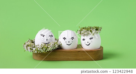 Three white eggs with a cheerful face with white wreath of spring flowers on a green background. Happy Easter greeting card. 123422948