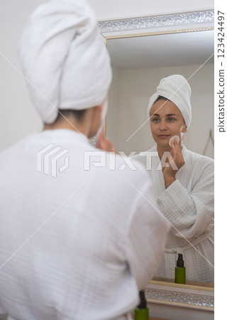 A woman, near the bathroom mirror, practices self-care by cleansing her skin with a cotton pad. A woman, near the bathroom mirror, practices self-care by cleansing her skin with a cotton pad. 123424497