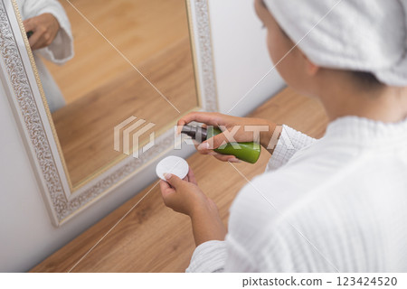 A woman, in her bathroom, attentively incorporates cleansing lotion into her skincare regimen using a cotton pad by the mirror.  123424520