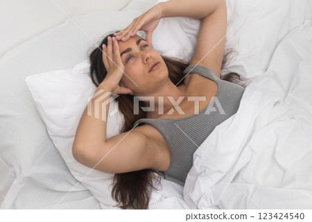 Frowning young lady, kneads temples, suffering a headache, reclined in a sunlit bed. Frowning young lady, kneads temples, suffering a headache, reclined in a sunlit bed. 123424540