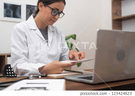 Medical Chart Examination: A female doctor in a white lab coat reviews patient medical documents while working at a table in a modern clinic.  123424623