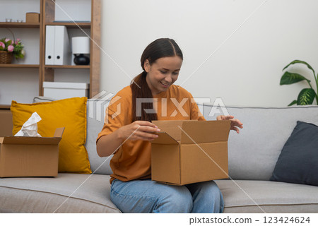 Smiling woman unboxing cardboard parcel ordered in internet while sitting on sofa female satisfying with good online shopping experience in modern apartment 123424624