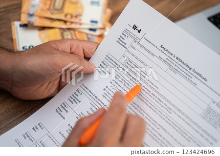 Close-up of a man's hands filling out a W-4 tax form with an orange pen, with euro banknotes in the background. Tax filing and financial management concept during tax season  123424696
