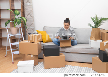 Smilling lady with bun o head with delighted expression sits on sofa collecting cardboard boxes for moving in new apartment with all modern furniture Smilling lady with bun o head with delighted expression sits on sofa collecting cardboard boxes for moving in new apartment with all modern furniture 123424792