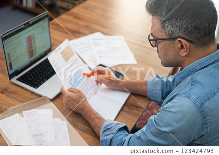Man reviewing utility bills at home with laptop and documents on wooden table. Concept of household expenses, family budget, and financial planning  Man reviewing utility bills at home with laptop and documents on wooden table. Concept of household expenses, family budget, and financial planning  123424793
