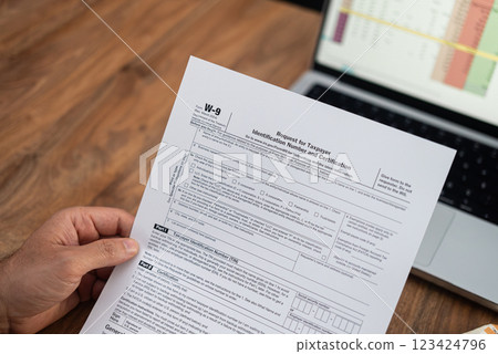 Close-up of a hand holding a W-9 tax form with a laptop in the background. Tax preparation and financial documentation concept, ideal for accounting and tax season themes Close-up of a hand holding a W-9 tax form with a laptop in the background. Tax preparation and financial documentation concept, ideal for accounting and tax season themes 123424796