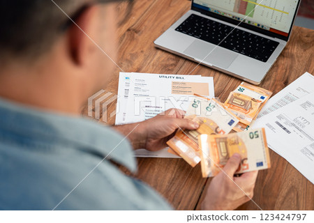 Man holding euro banknotes while reviewing utility bills and using laptop on wooden table. Concept of managing household finances and budgeting for monthly expenses Man holding euro banknotes while reviewing utility bills and using laptop on wooden table. Concept of managing household finances and budgeting for monthly expenses 123424797