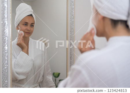 In a bathrobe, a woman enhances her radiance, applying skincare products and captivated by her reflection in the mirror. In a bathrobe, a woman enhances her radiance, applying skincare products and captivated by her reflection in the mirror. 123424813