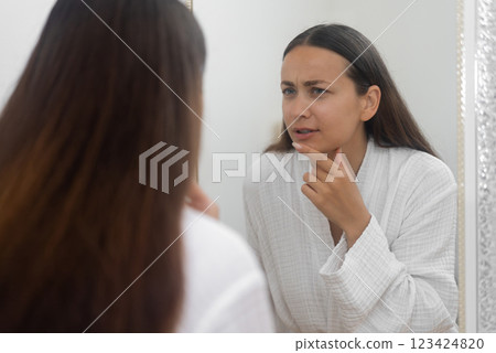 In the bathroom, a concerned woman looks at her reflection with sadness, distressed by her skin problems.  123424820