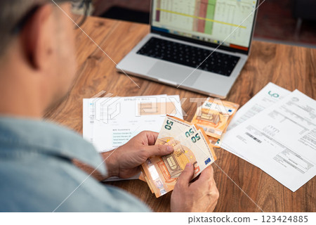 Old man counts euros banknotes sitting at table with unpaid utility bills and laptop, mature male customer plans budget for monthly household expenses in office, cash finances 123424885