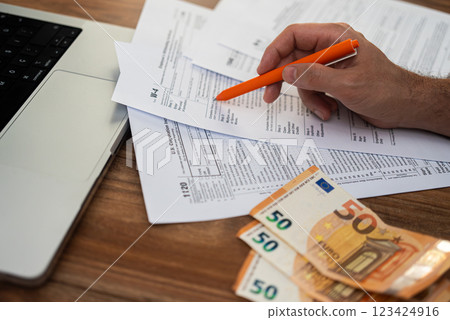 Office worker hand points with pen at tax forms near euro banknotes carefully examining steps of filling blank boxes correctly at wooden desk with modern laptop Office worker hand points with pen at tax forms near euro banknotes carefully examining steps of filling blank boxes correctly at wooden desk with modern laptop 123424916