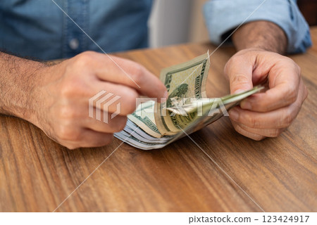 Male entrepreneur holds dollar banknotes in hands preparing money required for payment services of seasonal employees sitting at workplace in company office Male entrepreneur holds dollar banknotes in hands preparing money required for payment services of seasonal employees sitting at workplace in company office 123424917