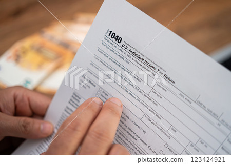 Male taxpayer holds tax form 1040 carefully examining requirements for filling Individual Income Tax Return blank sitting at workplace with banknotes in office Male taxpayer holds tax form 1040 carefully examining requirements for filling Individual Income Tax Return blank sitting at workplace with banknotes in office 123424921