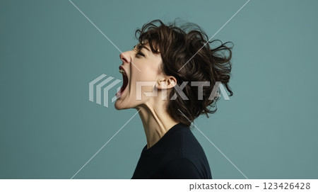 Profile of a young woman with wavy hair shouting with her mouth wide open and her head tilted back against a blue background, conveying strong emotions of anger, frustration, or protest 123426428