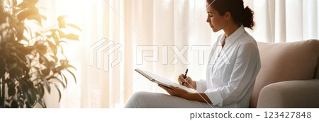 Young woman wearing a white shirt is sitting on a sofa near a window, bathed in sunlight, while writing in a notebook with a pen 123427848