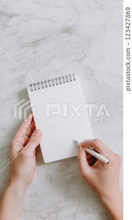 Woman is starting to write using a white pen on a blank spiral notebook placed on a white marble table, perfect for adding custom text or graphic Woman is starting to write using a white pen on a blank spiral notebook placed on a white marble table, perfect for adding custom text or graphic 123427869