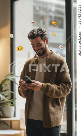 Happy young man is using mobile phone and smiling while social media icons and emojis float around his head, suggesting positive engagement and interaction online 123428212