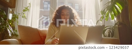 Comfortable young woman sitting on sofa using laptop at home during daylight, with houseplants in foreground and background and bright sunlight streaming through window 123429037