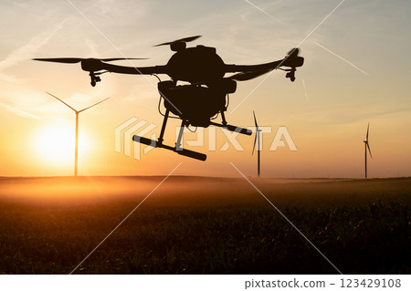 Drone sprayer flies over the agricultural field. Wind turbines on a horizon. 123429108