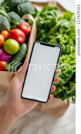 Hand holding a smartphone with blank white screen with a grocery bag full of fresh vegetables and fruits in background, promoting online shopping for groceries 123429161