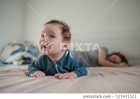 A three-month-old boy lies on his stomach on the bed and his mother sleeps behind him. Postpartum depression. A three-month-old boy lies on his stomach on the bed and his mother sleeps behind him. Postpartum depression. 123431321