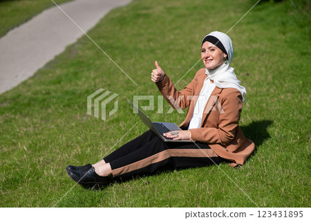 A young woman wearing a hijab sits on a lawn and uses a laptop outdoors and shows a thumbs up. A young woman wearing a hijab sits on a lawn and uses a laptop outdoors and shows a thumbs up. 123431895