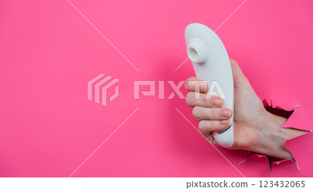 Female hand sticks out white vacuum clitoral vibrator through pink paper background.  123432065