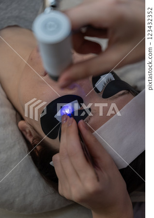A woman undergoing eyelash extension procedure using an ultraviolet lamp. A woman undergoing eyelash extension procedure using an ultraviolet lamp. 123432352