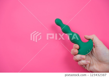 Female hand sticking out with green anal beads from pink paper background. Copy space. Female hand sticking out with green anal beads from pink paper background. Copy space. 123432387