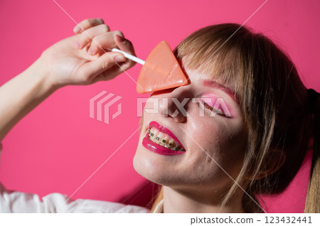 Portrait of a young woman with braces and bright makeup eating a lollipop on a pink background. 123432441