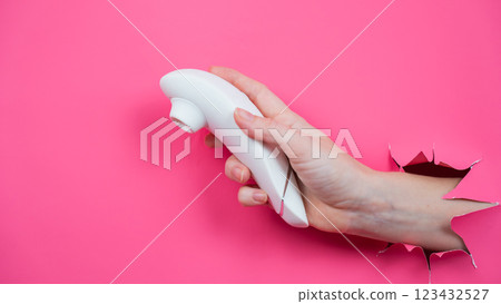 Female hand sticks out white vacuum clitoral vibrator through pink paper background.  123432527