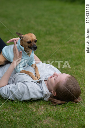Young Caucasian woman lying on the lawn while walking with a small dog. Vertical photo.  123433216
