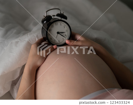 A pregnant woman lies in bed and holds an alarm clock. Close up of the belly. 123433324