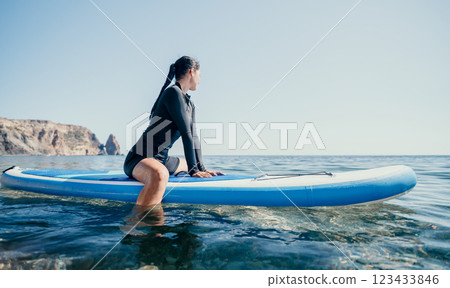 Woman Paddleboarding Ocean - A woman wearing a wetsuit is paddleboarding on a calm ocean with a rocky cliff in the background. Woman Paddleboarding Ocean - A woman wearing a wetsuit is paddleboarding on a calm ocean with a rocky cliff in the background. 123433846