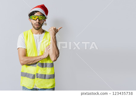 Amazed builder engineer in christmas hat pointing a copy space. Engineer man in christmas hat showing a product, Surprised Builder worker with christmas hat pointing a christmas promotion 123435738
